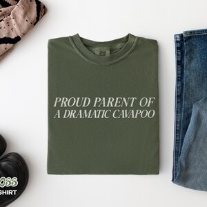 May include: An olive green crew neck t-shirt featuring the white text PROUD PARENT OF A DRAMATIC CAVAPOO printed in a clean, sans-serif font across the center of the chest. The garment has a ribbed collar and a standard short-sleeve construction.