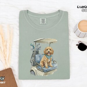 May include: Sage green t-shirt with a watercolor illustration of a golden doodle dog sitting in a golf cart. The dog wears a blue bandana. Golf clubs are in a bag behind the dog. The text "BAY T-SHIRT" is at the bottom left.