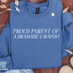 May include: A blue crewneck sweatshirt featuring the white text PROUD PARENT OF A DRAMATIC CAVAPOO printed across the chest. The garment has long sleeves, a ribbed collar, and a Gildan brand label visible at the inner neckline of the blue fabric.