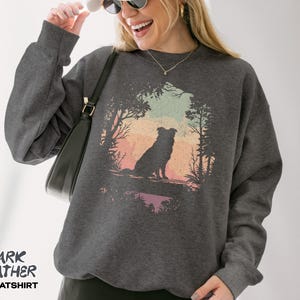 May include: Dark grey sweatshirt with a dog silhouette set against a colourful sunset scene. The text "DARK HEATHER SWEATSHIRT" is printed at the bottom. The sweatshirt has long sleeves and is made of a soft material.