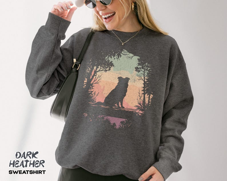 May include: Dark grey sweatshirt with a dog silhouette set against a colourful sunset scene. The text "DARK HEATHER SWEATSHIRT" is printed at the bottom. The sweatshirt has long sleeves and is made of a soft material.