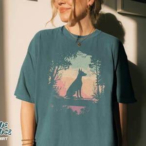 Pu&ograve; includere: T-shirt color verde acqua con una grafica di un cane silhouette in una scena di foresta. Il design presenta un cane seduto di fronte a un tramonto colorato, incorniciato da alberi. Il testo "BLUE SPRUCE T-SHIRT" &egrave; stampato in basso a sinistra.