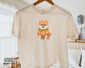 Può includere: Una t-shirt color avorio a maniche corte con un'illustrazione centrale in stile acquerello di un soffice cane Pomerania color marrone chiaro che indossa un costume da zucca arancione con foglie verdi intorno al collo. Girocollo e vestibilità ampia.