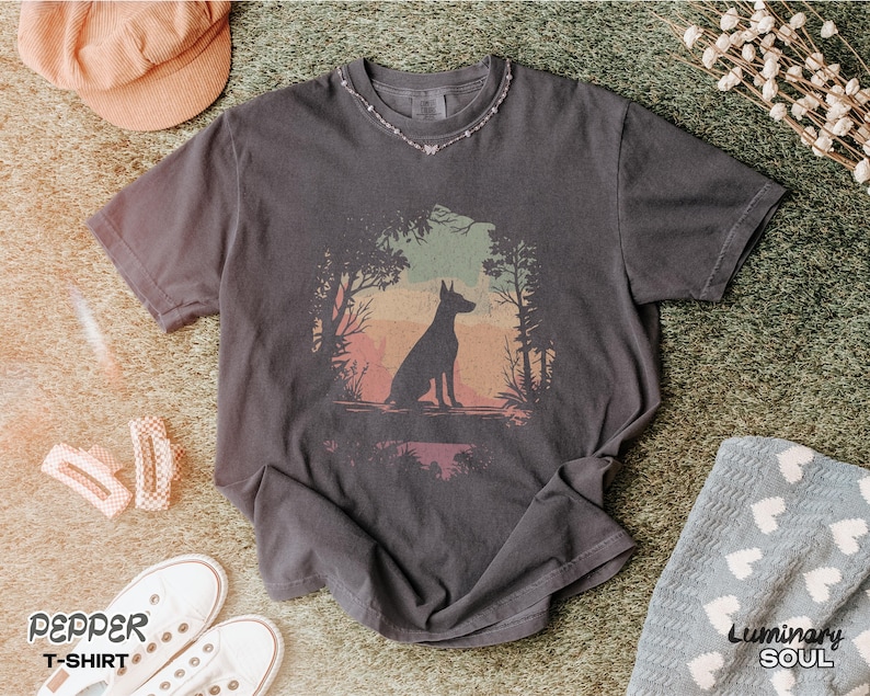 Pu&ograve; includere: T-shirt grigio scuro con una grafica di una silhouette di cane in una scena di foresta con un tramonto colorato. La maglietta &egrave; abbinata a scarpe da ginnastica bianche, un cappello rosa e fermagli per capelli. Il testo "Pepper T-Shirt" &egrave; visibile.