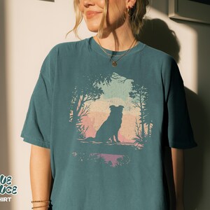 May include: Teal t-shirt with a dog silhouette in a forest scene against a sunset. The text "BLUE SPRUCE T-SHIRT" is at the bottom left. The design is framed by trees and foliage, creating a nature-inspired aesthetic.