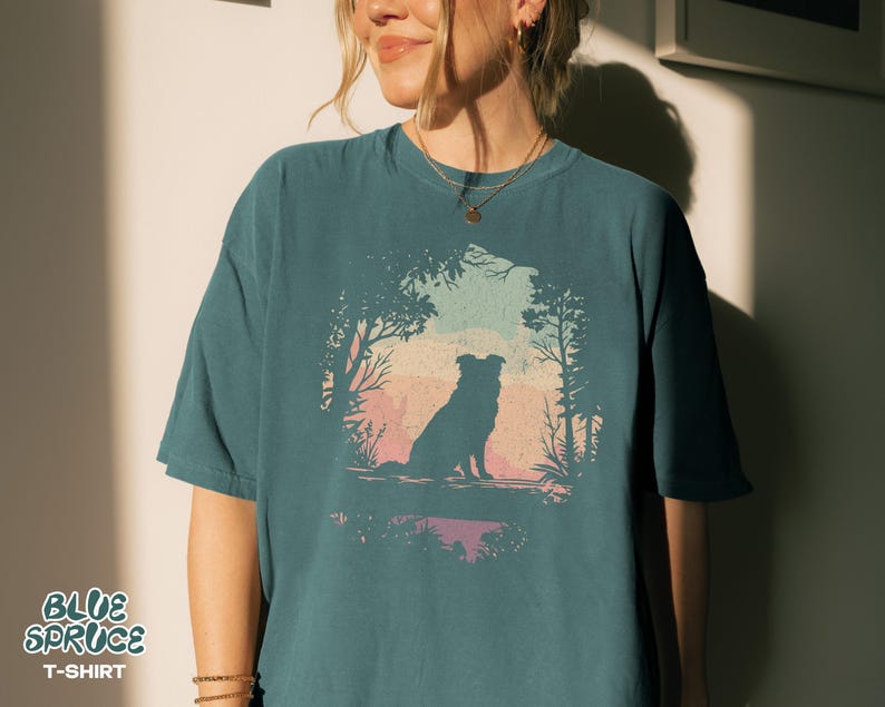 May include: Teal t-shirt with a dog silhouette in a forest scene against a sunset. The text "BLUE SPRUCE T-SHIRT" is at the bottom left. The design is framed by trees and foliage, creating a nature-inspired aesthetic.