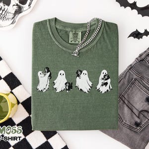May include: A moss green t-shirt featuring a graphic print of four white ghosts interacting with small black and white puppies. The shirt has a crew neckline and a Comfort Colors brand label at the inner collar. The design is centered on the chest.