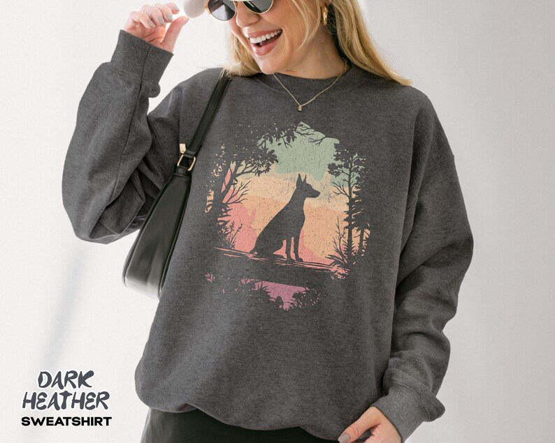 Pu&ograve; includere: Felpa grigio scuro con la silhouette di un cane in una scena di foresta con un colorato sfondo di tramonto. La felpa ha maniche lunghe e girocollo. Il testo "DARK HEATHER SWEATSHIRT" &egrave; stampato in basso.