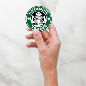 May include: A green and black round sticker with the text "KETAMINE VERSED" and a stylized image of a mermaid with a crown.