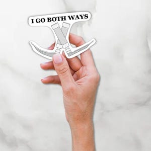 May include: A white sticker with the text "I GO BOTH WAYS" and a graphic of crossed tools. The sticker is held in a hand against a white and gray background. The tools appear to be a combination of a paint roller and a blade.