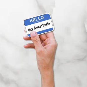 May include: A blue and white name tag with the text "HELLO" and "Hey Anesthesia". The name tag is held in a hand against a white marble background.