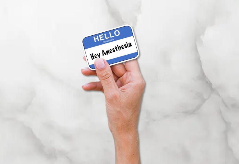 May include: A blue and white name tag with the text "HELLO" and "Hey Anesthesia". The name tag is held in a hand against a white marble background.