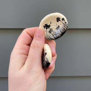 May include: Two small, oval ceramic dishes. Each dish has a black and white landscape design with stylized plants and grasses. The dishes are held against a grey background.