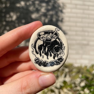Puede incluir: Pin de cerámica ovalado con una ilustración en blanco y negro de un caldero sobre llamas. El caldero contiene un hueso y líquido burbujeante. El pin se sostiene contra un fondo borroso.