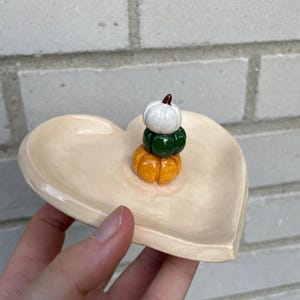 Könnte beinhalten: Eine herzförmige Keramikschale mit einem Stapel von drei Mini-Kürbissen. Die Kürbisse sind in Weiß, Grün und Orange bemalt.