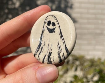 May include: A round, off-white ceramic button featuring a hand-drawn ghost design. The ghost has a smiling face and flowing body, rendered in black ink. The button is held against a blurred background of green foliage and sunlight.
