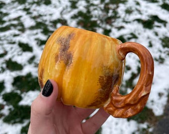 May include: A yellow ceramic pumpkin mug with a brown handle. The mug is shaped like a pumpkin and has a brown glaze.