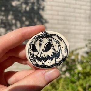 May include: A round, off-white ceramic button with a black, hand-drawn jack-o'-lantern face. The pumpkin has triangular eyes, a jagged mouth, and a stem. The button is held in a person's hand against a blurred background.