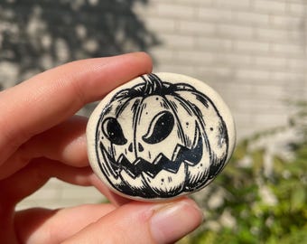 May include: A round, off-white ceramic button with a black, hand-drawn jack-o'-lantern face. The pumpkin has triangular eyes, a jagged mouth, and a stem. The button is held in a person's hand against a blurred background.