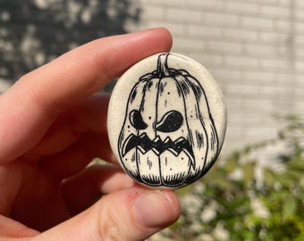 May include: A round, off-white button featuring a black, hand-drawn jack-o'-lantern face. The pumpkin has a carved, angry expression with jagged teeth and a stem. The button is held against a blurred green and white background.