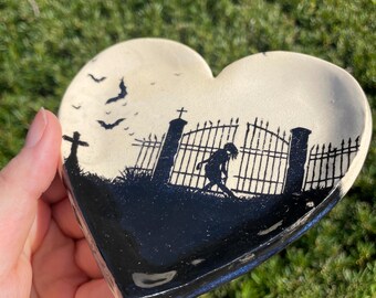 May include: A heart-shaped ceramic dish with a black and white design of a graveyard scene. The scene includes a gate, a cross, and a figure walking through the gate. There are bats flying in the sky.