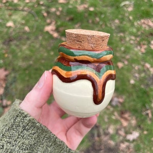 May include: A small, round, white ceramic jar with a cork lid. The jar is decorated with brown, green, and orange stripes. The stripes are painted to look like they are dripping down the side of the jar.
