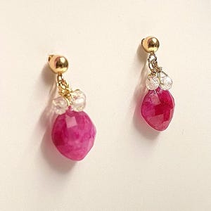 Puede incluir: Un par de pendientes dorados con una gema rosa intenso facetada y colgante. Los pendientes también presentan pequeñas piedras de acento claras y redondas, sobre un fondo blanco.