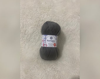 May include: A skein of dark gray yarn with a white paper label featuring the brand name Kartopu, the product name Baby One, and the text 100g / 250m. The label also displays small illustrations of baby clothing and accessories.