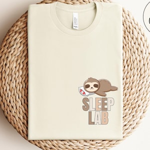 May include: A light beige t-shirt with a cartoon sloth sleeping on a pillow and the text "SLEEP LAB" printed on the front. The shirt is on a woven straw surface.