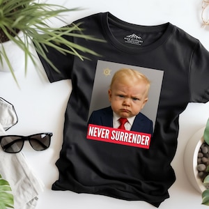 May include: Black t-shirt featuring a baby with a stern expression, wearing a suit and tie, with the text "NEVER SURRENDER" in a red banner. The shirt has a crew neck and short sleeves.