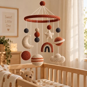 May include: A hanging nursery mobile featuring crocheted celestial and weather-themed elements in rust, cream, and dark gray. The mobile includes a crescent moon, a star, a rainbow, a cloud, and spherical planets suspended from a circular frame.