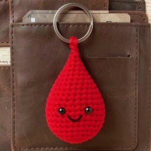 May include: A red crochet keychain shaped like a teardrop with two small black bead eyes and a stitched black smile. The keychain features a silver metal ring attached to the top of the red yarn figure, which has a textured, knotted surface.