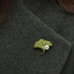 Può includere: Spilla a forma di foglia di ginkgo verde con un accento di perla. La spilla è in metallo e presenta un design dettagliato e strutturato. La spilla è appuntata su un cappotto grigio scuro.