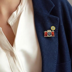 May include: A gold-toned camera pin with red and black accents, featuring a yellow flash. The pin is attached to a navy blue blazer, worn over a cream-colored blouse. The camera pin is a unique accessory.