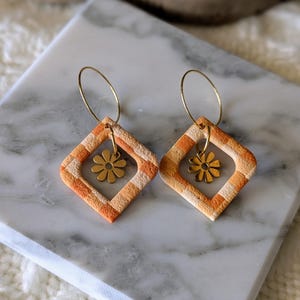 Könnte beinhalten: Ein Paar handgefertigte Ohrringe mit geometrischem Design. Jeder Ohrring hat einen rautenförmigen Anhänger mit orangefarbenen und cremefarbenen Streifen, einen goldfarbenen Blumenanhänger und einen goldenen Reifen. Die Ohrringe werden auf einer Marmoroberfläche präsentiert.