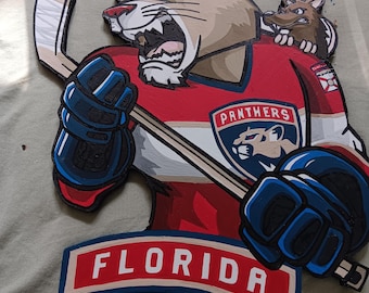 May include: Wooden sign featuring a cartoon cougar in a hockey uniform, holding a hockey stick. The sign is in the shape of the cougar and has the word "FLORIDA" at the bottom. The cougar is wearing a red, white, and blue uniform.