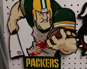 May include: A wooden wall hanging featuring a cartoon football player wearing a green and yellow helmet and jersey, holding a brown football in one hand and a white meat cleaver in the other, with red blood splatters and the text PACKERS.