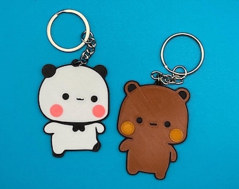 May include: Two keychains with cute animal designs. One keychain features a black and white panda bear with pink cheeks and a black bow tie. The other keychain features a brown bear with orange cheeks. Both keychains have silver key rings.