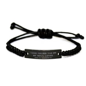 Puede incluir: Pulsera trenzada negra con una placa metálica rectangular. La placa tiene la inscripción "I HAVE FOUND THE ONE MY SOUL LOVES. SONG OF SOLOMON 3:4" en letras blancas. La pulsera es ajustable.