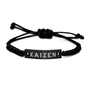 May include: Black adjustable bracelet with a rectangular plate inscribed with "KAIZEN" in white. The bracelet is made of a woven black cord and has a sliding knot fastening. A simple, minimalist design.