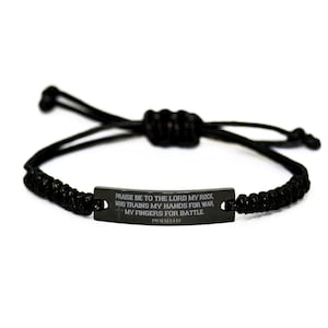 Pode incluir: Pulseira trançada preta com uma placa de metal retangular. A placa apresenta o texto gravado: "PRAISE BE TO THE LORD MY ROCK, WHO TRAINS MY HANDS FOR WAR, MY FINGERS FOR BATTLE." O texto está em uma fonte sem serifa. A pulseira é ajustável.