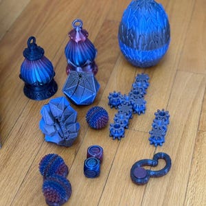 Könnte beinhalten: Eine Sammlung von 3D-gedruckten Objekten in blau-violetten Farbverläufen, darunter zwei laternenförmige Ornamente, ein texturiertes Ei, eine geometrische Polyederform, ein Kristallcluster, zwei stachelige Kugeln, ein Zahnradkreuz, ein Zahnradstreifen, zwei Würfel und eine Schlaufe.