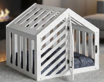 Puede incluir: Una jaula de madera blanca para mascotas con techo a dos aguas y paredes de listones. La estructura cuenta con una puerta abatible con pestillo de metal negro y un cojín acolchado azul oscuro en el interior, con listones diagonales al frente.