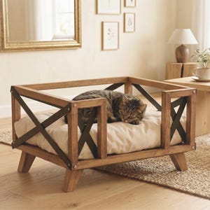 May include: A rectangular pet bed featuring a light brown wooden frame with dark metal X-shaped accents on the sides. A soft, cream-colored cushion rests inside the frame, providing a comfortable surface for a sleeping brown tabby cat.