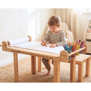 Op de afbeelding: Een lichtgekleurde houten activiteitentafel met een ingebouwde papierrolhouder aan één kant en drie cilindervormige mesh potloodhouders aan de hoek, gevuld met diverse kleurpotloden om op het witte papieroppervlak te tekenen.