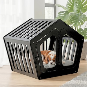 May include: A black, house-shaped pet crate with a slatted roof and side panels. The structure features a front opening and contains a gray, cushioned mat inside. A brown and white dog is resting comfortably on the mat within the enclosure.