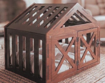 Puede incluir: Una casa para mascotas de madera marrón oscuro con techo de listones y una entrada frontal con dos puertas abatibles con marco en forma de X. Dentro de la estructura, se ve un cojín de color claro a través de los listones laterales.