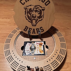May include: Wooden cribbage board with a Chicago Bears logo and text. The board has a circular design with scoring holes, pegs, and a card holder. A deck of cards is visible. The top piece has the words "Chicago Bears" and a bear logo.