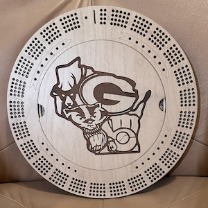 May include: Circular wooden cribbage board with a laser-etched design featuring a badger and the letter "G". The board has numerous holes for pegs and is marked with numbers for scoring. The board is made of light-colored wood.