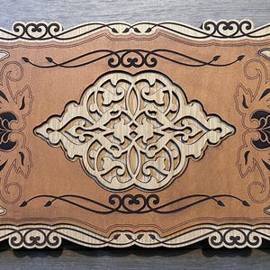 May include: A rectangular wooden wall plaque featuring intricate laser-cut layered designs. The piece displays a central symmetrical filigree pattern surrounded by ornate corner motifs and a decorative scalloped border in light and dark wood tones.